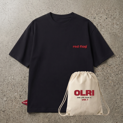 A black short-sleeve t-shirt with red 'red flag' text on the chest, laid flat on a textured gray concrete background next to a cream-colored drawstring bag printed with 'OLRI'.