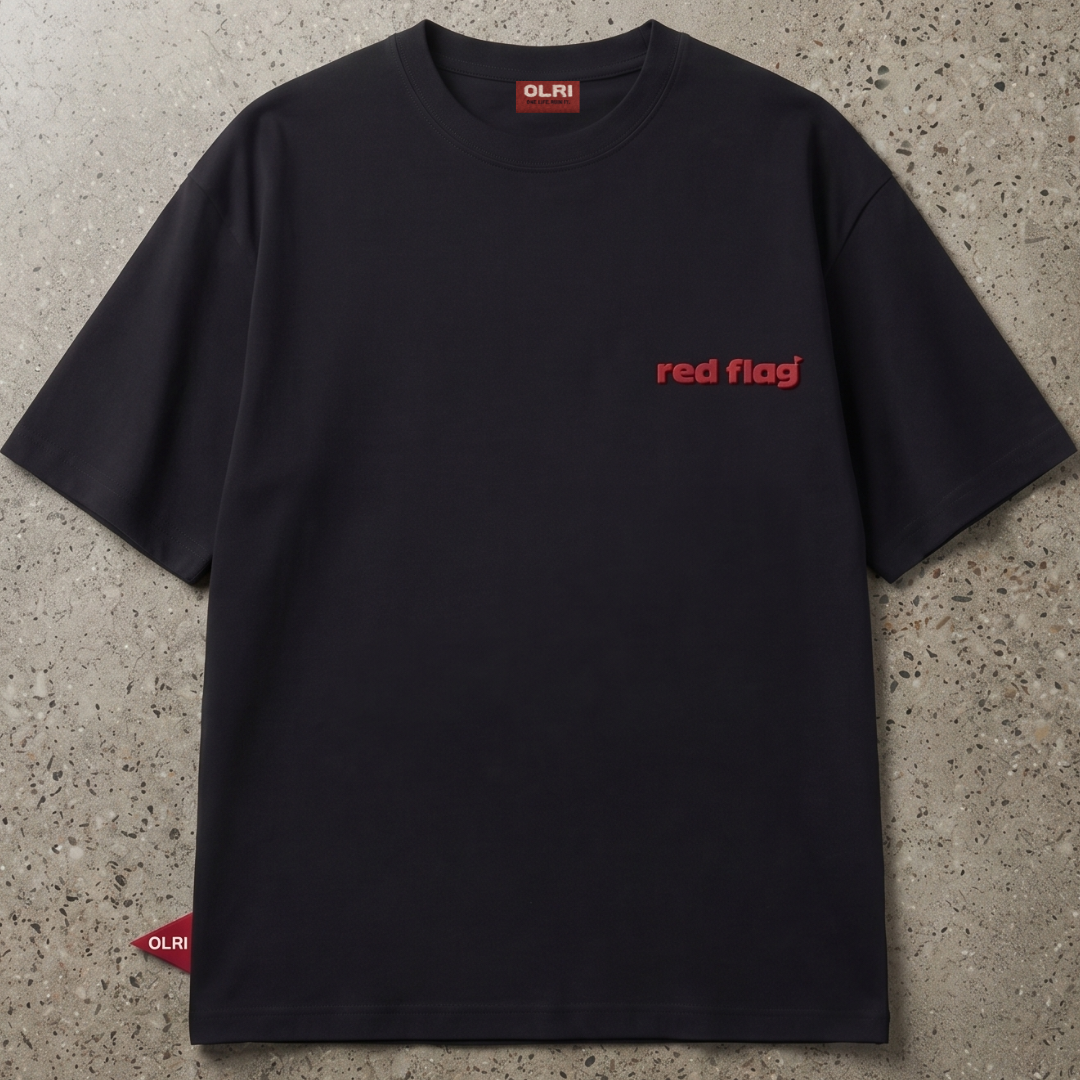 A black short-sleeve crewneck t-shirt with 'red flag' printed in red on the left chest, laid flat on a textured gray concrete surface. The brand 'OLRI' is visible on the inner neck label and a small red tag near the bottom left hem.