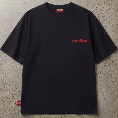 A black short-sleeve crewneck t-shirt with 'red flag' printed in red on the left chest, laid flat on a textured gray concrete surface. The brand 'OLRI' is visible on the inner neck label and a small red tag near the bottom left hem.