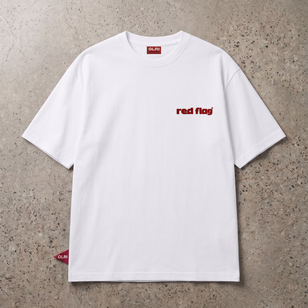 A white short-sleeve crewneck t-shirt with 'red flag' printed in red on the left chest, laid flat on a textured concrete surface.